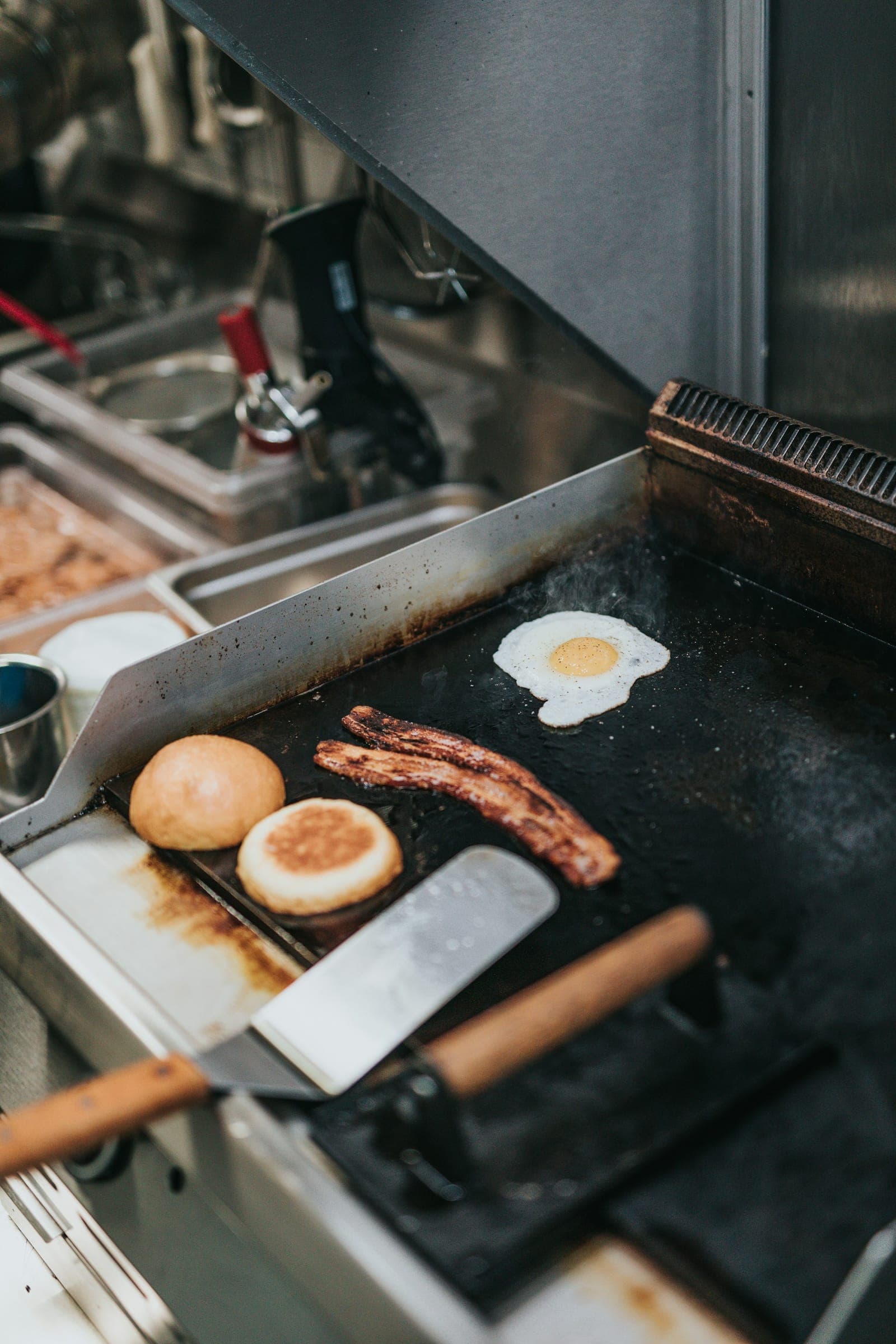Breakfast of eggs, bacon, and buns cooking on a flat-top griddle